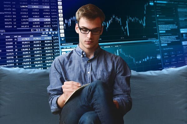 joven con gafas escribiendo en un cuaderno. Al fondo gráficas de mercado de valores. Colores azules. Foto alusiva al desarrollo empresarial